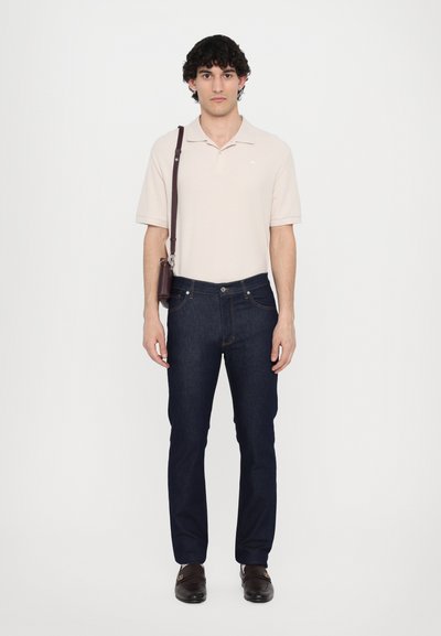 Cămașă polo bej, blugi negri, și mocasini maro; prezintă o geantă de umăr și un material ușor într-o croială lejeră. Design simplu, fără modele.