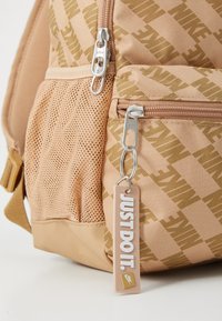 Ferskenfarget nylon ryggsekk med gullfarget Nike-logo mønster, nettinglomme på siden, sølvfargede glidelåser, og et beige merkelapp som står "JUST DO IT."
