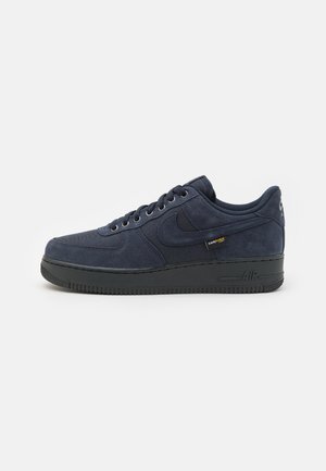Granatowy zamszowy sneaker Nike Air Force 1 z czarną podeszwą, sznurowadłami i metką z tkaniny Cordura z boku.