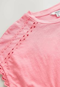 Camiseta de algodón rosa con cuello redondo y bordes en forma de ondas, con detalles recortados en las mangas. Textura suave, tejido ligero.