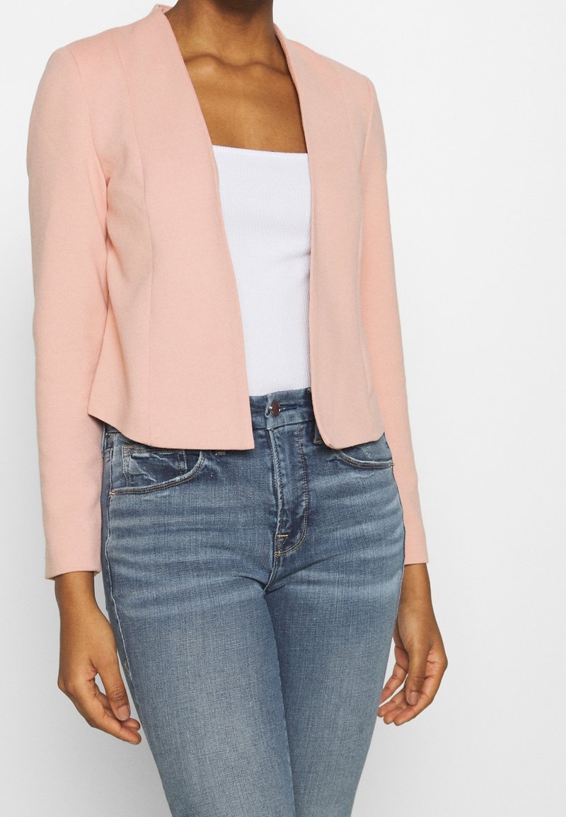 Femme portant un blazer rose clair ouvert sur un haut blanc et un jean bleu taille haute sur fond blanc.