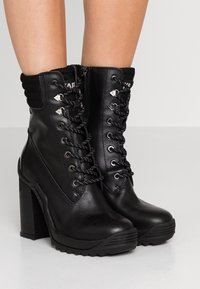 Botas negras de tobillo con un acabado de cuero liso, que presentan un frente con cordones, tacones altos y gruesos, y un cuello de punto texturizado.