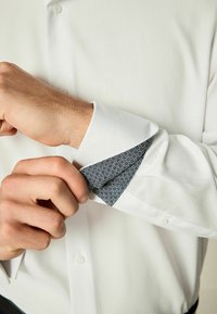 Vit skjorta med texturerad tyg. Manschetten visar ett mönstrat blått inre. Närbild av hand som justerar manschetten. Knappdetalj synlig.