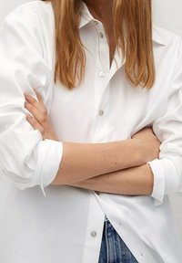 Biała koszula z guzikami, z podwiniętymi rękawami, o gładkiej fakturze, klasycznym kołnierzyku i małych guzikach. Nosi się ją na dżinsach.