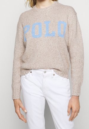 Beige knitted sweater with blue "POLO" text worn with high-waisted white pants, hands relaxed by sides, cropped at neck and thighs.