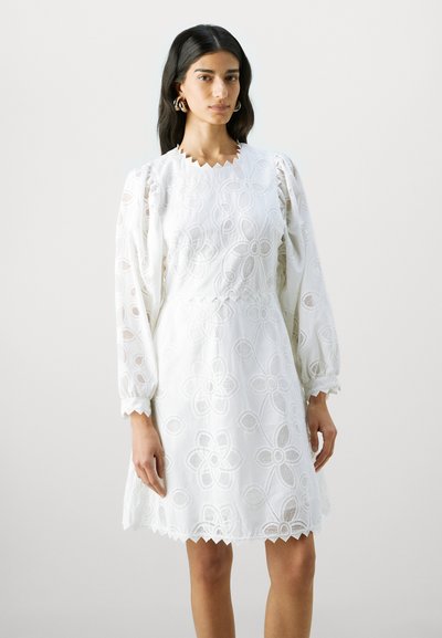 Robe blanche brodée ornée de motifs floraux, avec des manches longues bouffantes et un col et un ourlet à volants. Fabriquée en tissu léger.