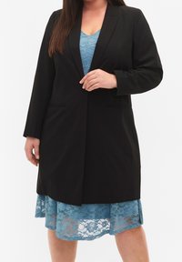 Blazer noir cintré à manches longues, avec deux poches avant. En dessous, une robe en dentelle bleu clair avec un ourlet fluide.