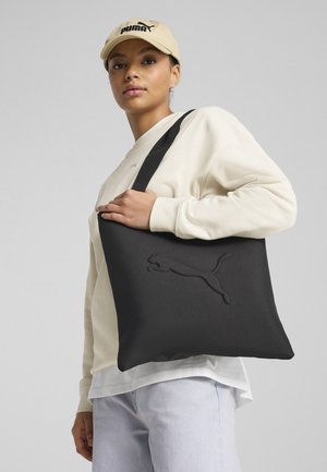 Czarna torba na ramię wykonana z miękkiego materiału, z wytłoczonym logo Puma. Zawiera czarny pasek i jest zestawiona z kremowym swetrem.