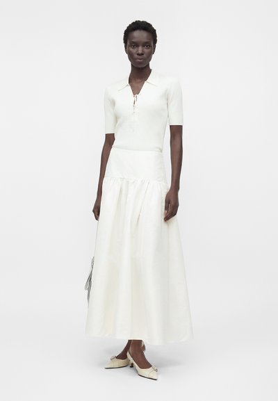 Femme portant un haut en maille blanc à manches courtes avec un col lacé et une longue jupe blanche taille haute avec des talons pointus couleur crème.