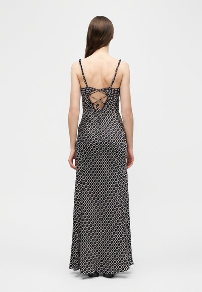 Γυναίκα με μακριά καστανή τρίχα, που φοράει μακρύ φόρεμα σε ασπρόμαυρο μοτίβο με λεπτές τιράντες και λεπτομέρεια κορδονιών που διασταυρώνονται στην πλάτη.