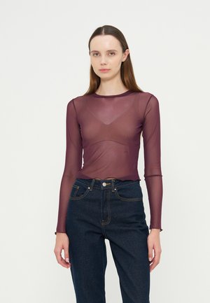 Jeune femme aux cheveux bruns raides porte un haut violet transparent à manches longues par-dessus un soutien-gorge et un jean taille haute bleu foncé sur un fond uni.