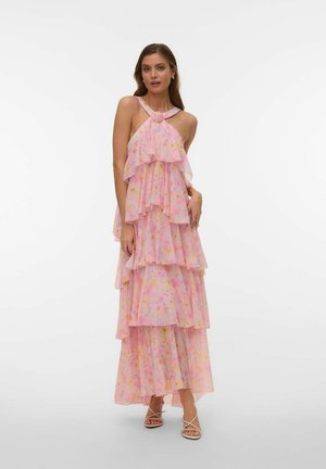 Vrouw in een mouwloze, gelaagde roze maxi-jurk met bloemenprint en een halternek met knoop, staand met één hand op de heup, draagt witte sandalen met bandjes.
