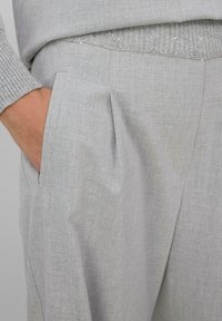 Pantalons châssis gris clair avec une texture lisse, dotés de poches latérales et de plis subtils, associés à un haut en tricot gris.