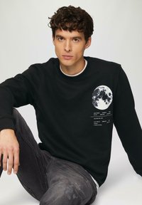 Zwarte sweatshirt met een ronde maanafbeelding en witte tekstdetails. Het model zit en draagt een paar grijze versleten jeans. Gladde stofstructuur.