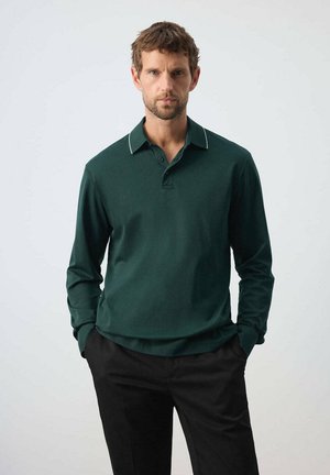 Hombre con pelo corto y castaño y barba, que lleva una camisa polo de manga larga verde oscuro y pantalones negros, de pie con las manos en los bolsillos.