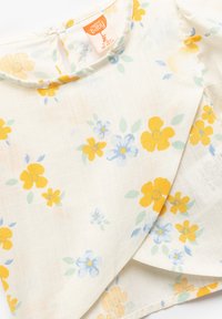 Top floral para bebés en tejido ligero de color crema con patrones de flores amarillas y azules. Presenta un escote redondo y un diseño superpuesto.