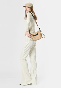 Gorra beige, mono largo crema con piernas acampanadas. Bolso bandolera marrón con etiqueta negra. La modelo está de perfil, mostrando los detalles del conjunto.