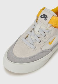 Ljusgrå mockaskor med gula accenter, grå tåskydd, vit gummisål, snördesign och 'Nike SB'-logotyp på tungan.