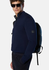 Maglione zip con zip di colore blu navale, con colletto alto e finitura testurizzata, abbinato a pantaloni scuri e uno zainetto nero con spallacci regolabili.
