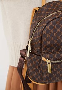 Zaino marrone con tessuto a motivi, hardware dorato e dettagli beige. Presenta cerniere e spallacci regolabili.