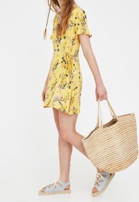 Robe portefeuille fleurie jaune à manches courtes, avec un lien à la taille. Associée à des espadrilles bleu clair et un sac fourre-tout en paille tressée.