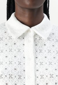 Camicia bianca in pizzo con colletto a punta, chiusura con bottoni, motivi intricati e superficie testurizzata.