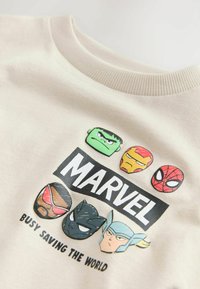 Bež trenirka z liki Marvela: Hulk, Iron Man, Spiderman, Black Panther in Thor, z drzno napisano znamko "MARVEL" in besedilom pod njo.