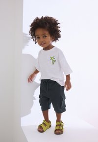 Enfant portant un T-shirt blanc avec un motif de palmier vert, un short cargo foncé et des sandales vertes à boucles, debout dans un décor minimaliste.