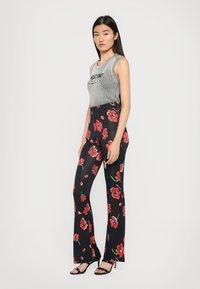 Kvinne står iført en grå ermeløs ribbestrikket topp og svarte høye bukser med røde blomsterprint og svarte hælsandaler.