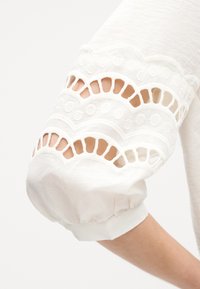 Bílý látkový rukáv s vroubkovanými krajkovými výřezy a vyšívanými kruhovými vzory, shrnutý u manžety na paži osoby.