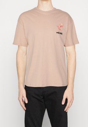 Homme portant un t-shirt à manches courtes rose clair avec un petit motif floral rouge et un texte noir sur le côté gauche de la poitrine, associé à un pantalon noir.
