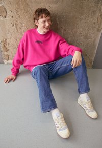 Sudadera rosa con texto negro, jeans azules y zapatillas crema con acentos y rayas en tonos pastel. Posición sentada contra una pared texturizada.
