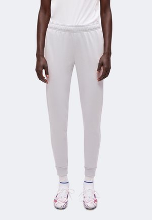 Personne portant un pantalon de jogging gris clair, des chaussures de sport blanches avec des détails rouges et bleus, et des chaussettes blanches avec des rayures bleues.