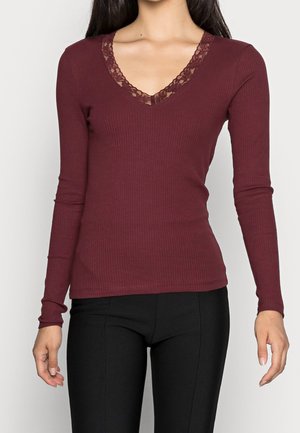 Kobieta ubrana w dopasowaną, długorękawową bordową bluzkę prążkowaną z dekoltem w kształcie litery V wykończonym koronką, zestawioną z czarnymi, wąskimi spodniami.