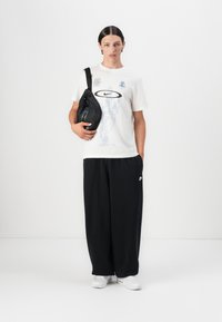 White t-shirt with blue graphic design, black sweatpants, and a black sling bag. Features Nike logo and relaxed fit. White sneakers complete the look.