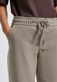 Beige trekbroek met zijstreep, elastische tailleband en hand in zak, gedragen met een bruine top.