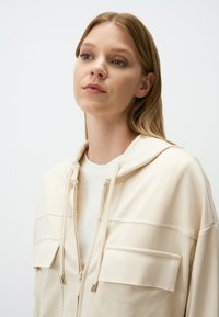 Crème-kleurige, ritssluiting hoodie met voorzakken, een trekkoordcapuchon en een relaxte pasvorm. Zacht, glad materiaal met een minimalistisch ontwerp.