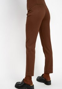 Osoba w dopasowanych brązowych spodniach, w brązowych skarpetach i czarnych, wsuwanych butach na platformie, stojąca na tle białym.