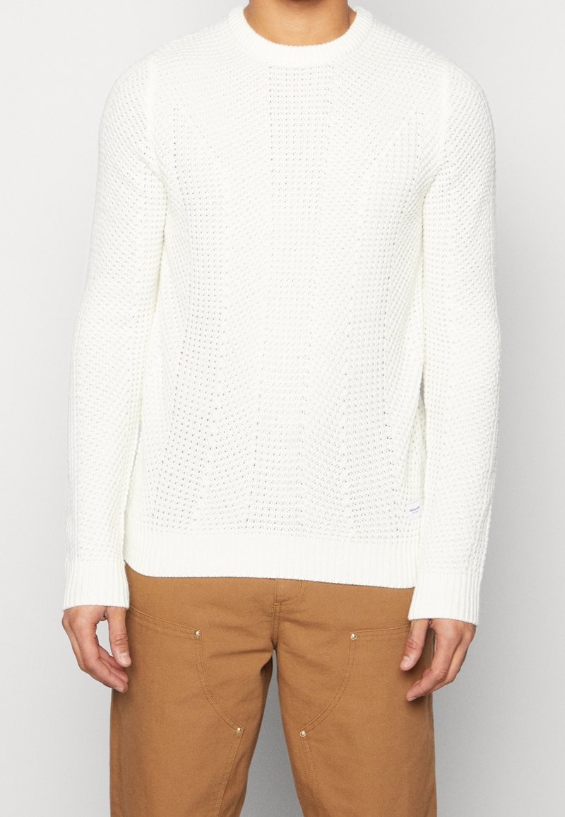 Witte gebreide trui met een gestructureerd patroon, ronde halslijn en geribde manchetten, gecombineerd met een paar bruine broeken.