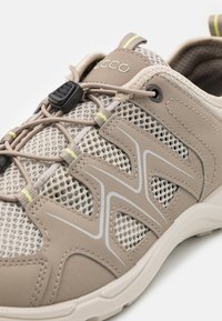 Scarpa sportiva beige chiaro realizzata in materiali in rete e sintetici lisci. Presenta un sistema di allacciatura elastico e dettagli testurizzati con elementi riflettenti.