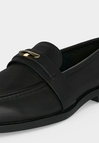 Mocassin en cuir noir à bout arrondi avec détail en métal doré incurvé sur la bride traversant le dessus.
