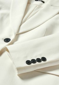 Crème blazer met een gladde textuur, voorzien van een ingezette reverskraag, zwarte knopen en decoratieve stiksels op de mouw met vier zwarte knopen.