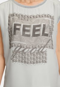 Chemise gris clair avec un graphisme texturé portant les mots "FEEL good" en gris foncé, rehaussée de clous et de motifs floraux.
