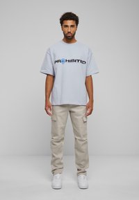 Lichtblauw T-shirt met korte mouwen met zwarte "PROHIBITED" grafiek en wereldbolpictogram, gecombineerd met beige cargobroek en witte sneakers.