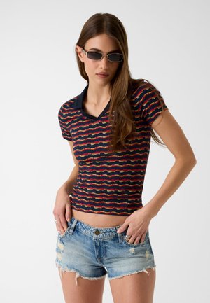 Kobieta w czarnych okularach przeciwsłonecznych, granatowej koszulce polo z czerwonymi i żółtymi falistymi paskami oraz przetartymi jasnoniebieskimi jeansowymi szortach.