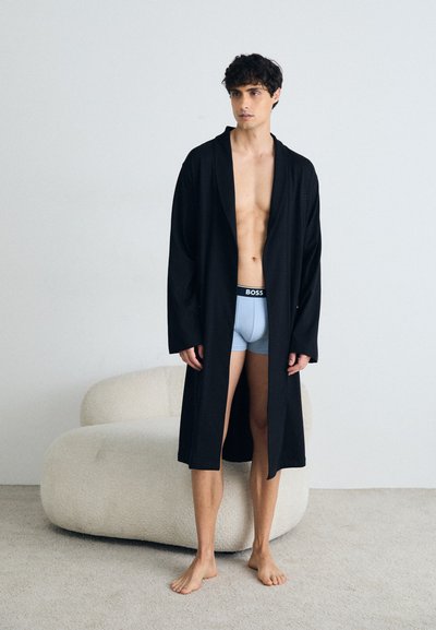 Une longue robe noire en tissu lisse, présentant une coupe ample et de larges manches, portée par-dessus un short de bain bleu clair, avec un canapé beige doux en arrière-plan.