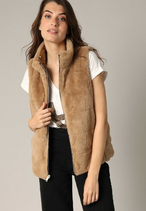 Veste sans manches en fausse fourrure beige avec un col haut et une fermeture éclair à l'avant, présentant un motif matelassé et des poches latérales, portée sur un t-shirt blanc.