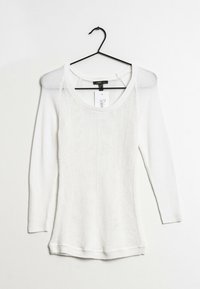 Maglietta bianca a maniche lunghe in maglia con scollo a barchetta, motivo testurizzato e orlo a coste. Appesa a un gancio di plastica nero su uno sfondo neutro.