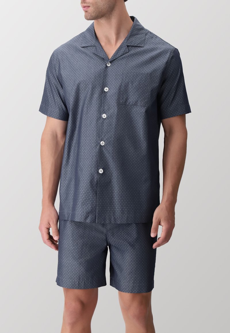 Pigiama blu navy a maniche corte con camicia bottonata dotata di tasca e motivo a pois bianchi; abbinato a pantaloni corti coordinati.