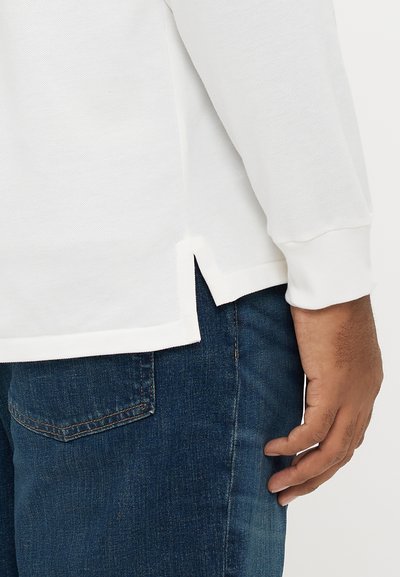 Λευκό τοπ μακρυμάνικο με υφή στο ύφασμα, πλαϊνές χαρακιές στο κατώφλι και ριμπ μανσέτες, σε συνδυασμό με σκούρο τζιν παντελόνι.